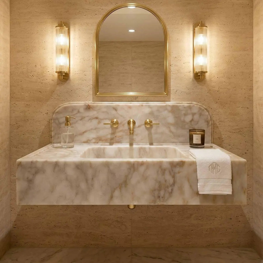 A luxury bathroom vanity featuring a floating marble sink with a white and beige veined pattern, gold fixtures, a matching marble backsplash, a gold-framed arched mirror, and two illuminated wall sconces. A folded white towel with a gold monogram is draped over the right side of the counter.