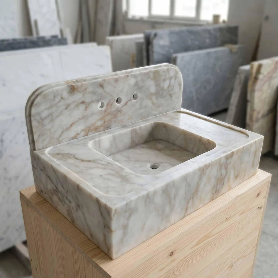 A rectangular marble bathroom sink with a matching backplate, displayed on a wooden pedestal in a showroom. The sink features a warm off-white base with natural beige and light brown veining.