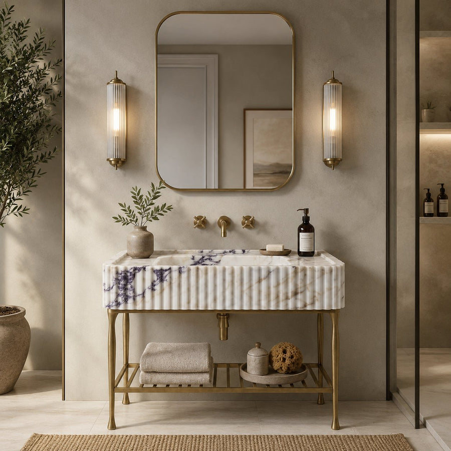 A modern bathroom vanity with a ribbed white marble countertop featuring subtle purple veining, supported by a gold metal frame. The vanity is styled with a gold faucet, gold wall sconces, a gold-framed mirror, and a gold shelf holding folded towels and decorative items.
