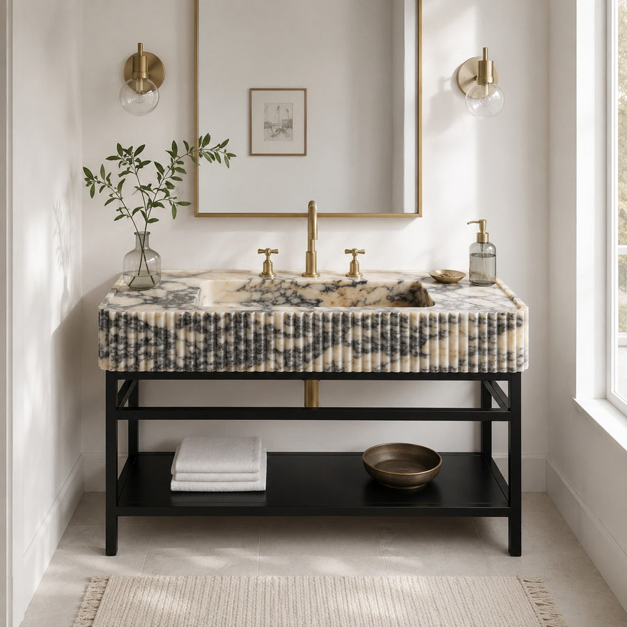 A modern bathroom vanity with a black metal frame and a marble-look top featuring a pleated front panel. The sink has a matte brass faucet and two separate hot/cold handles.