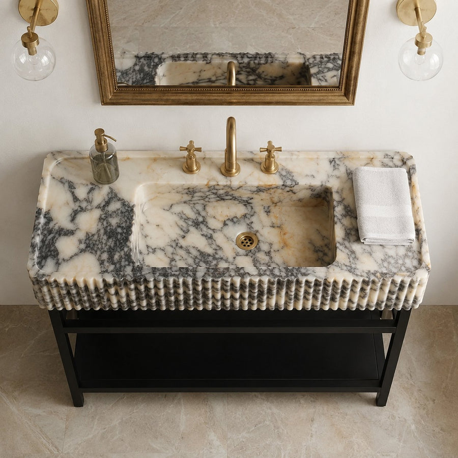 A luxury bathroom vanity featuring a rectangular carved marble sink with dramatic black and cream veining, mounted on a black metal frame. The sink is styled with a brass faucet, gold soap dispenser, and a folded white towel, set against a warm beige wall with a matching marble mirror.