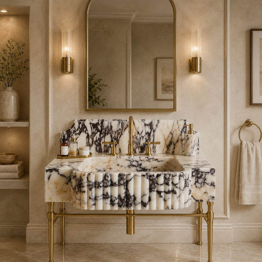 A luxury bathroom vanity featuring a white marble sink and matching marble backsplash with dramatic black veining, supported by ornate gold metal legs. The vanity is styled with gold fixtures, a matching marble soap dispenser, and a large gold-framed mirror on a beige paneled wall.