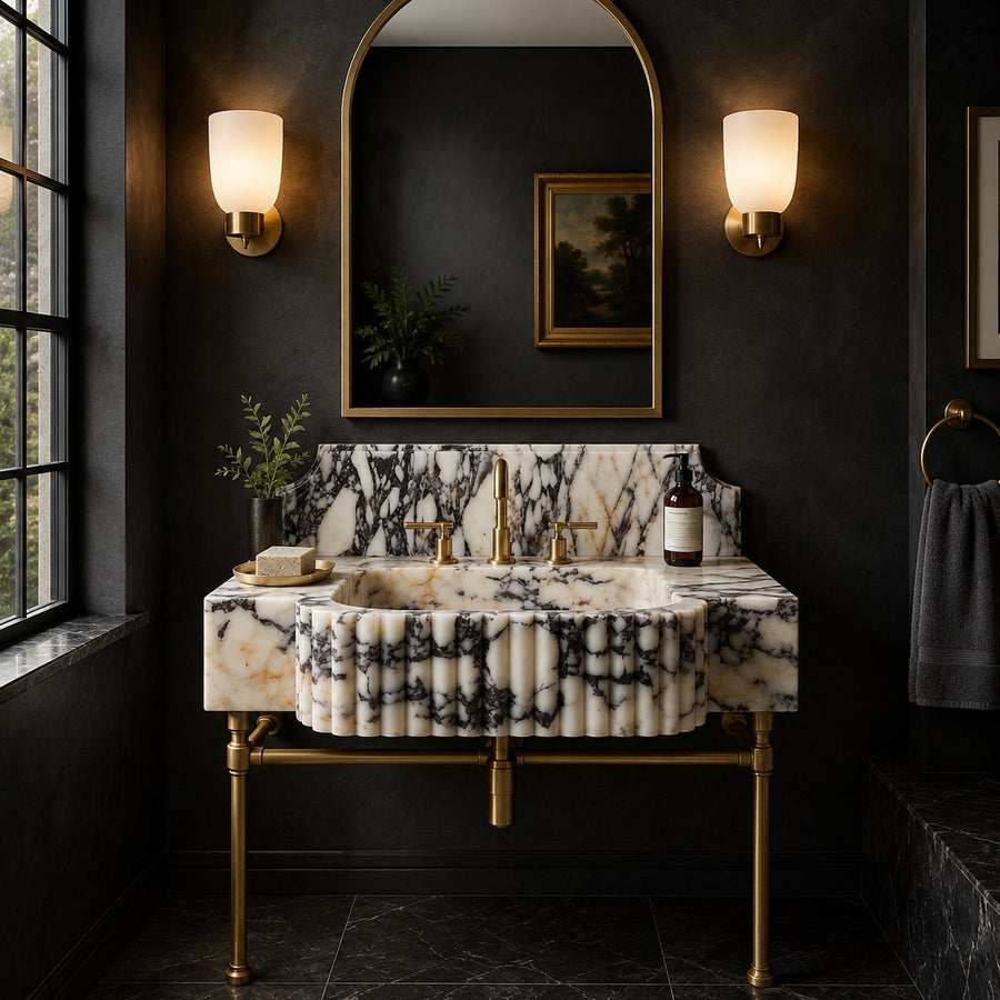 A luxury bathroom vanity featuring a ribbed white marble sink and matching marble backsplash with dramatic black veining, supported by brass legs and fixtures. The vanity is set against a dark charcoal wall with a large arched gold-framed mirror, flanked by two glowing wall sconces.