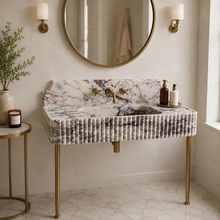 A modern vanity sink with a white and gray marble-look front, fluted sides, and gold-tone legs and faucet hardware, shown in a styled bathroom setting with a round mirror, wall sconces, and a side table.