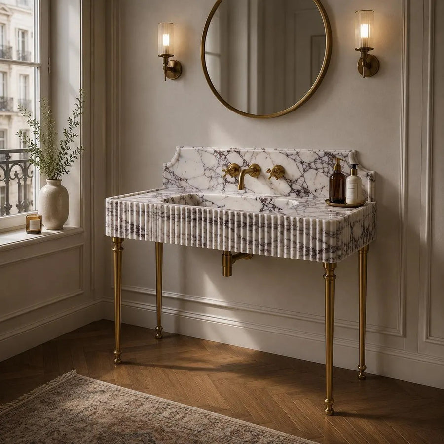 A freestanding bathroom vanity with a white marble sink and backsplash, fluted gold legs, and brass fixtures, styled in a classic cream-colored bathroom with a round mirror and wall sconces.