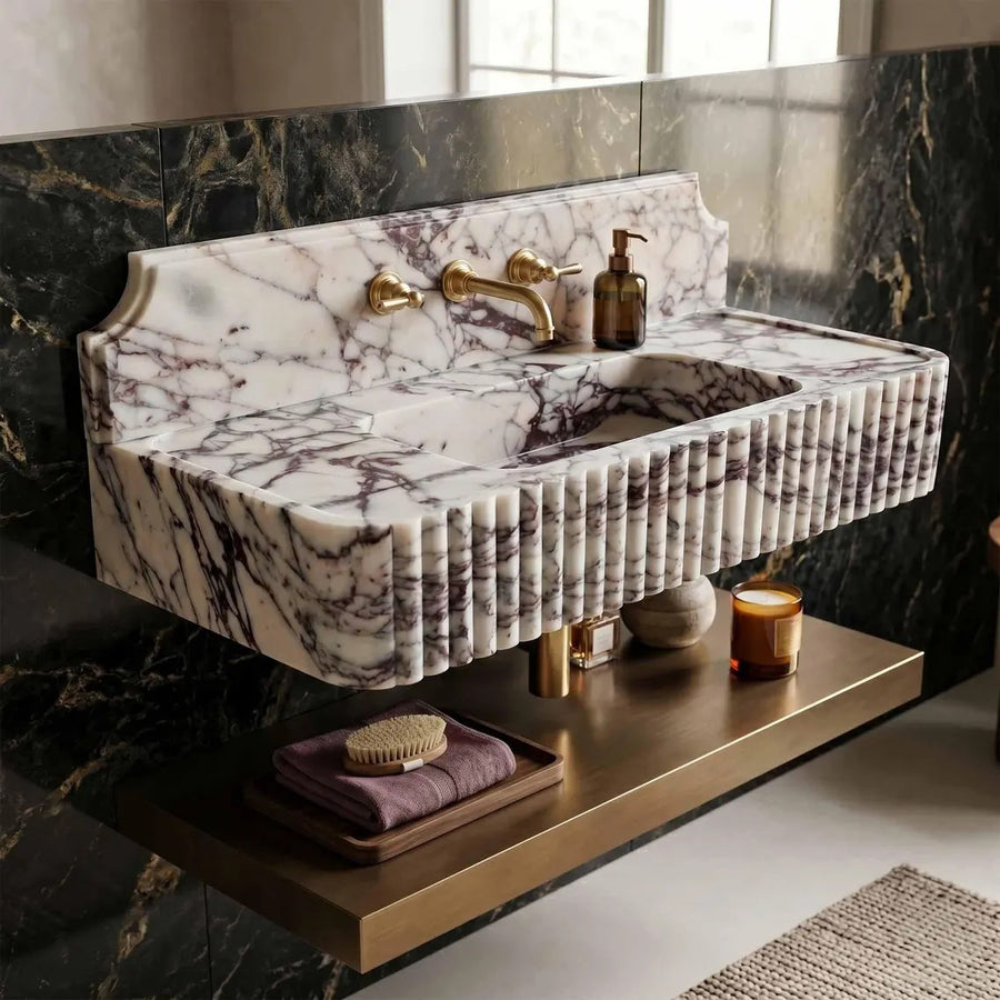 A floating bathroom sink vanity made of white marble with dark purple veining, mounted above a brushed brass floating shelf. The sink features a fluted front edge, a matching marble backsplash, and a brass faucet and soap dispenser.