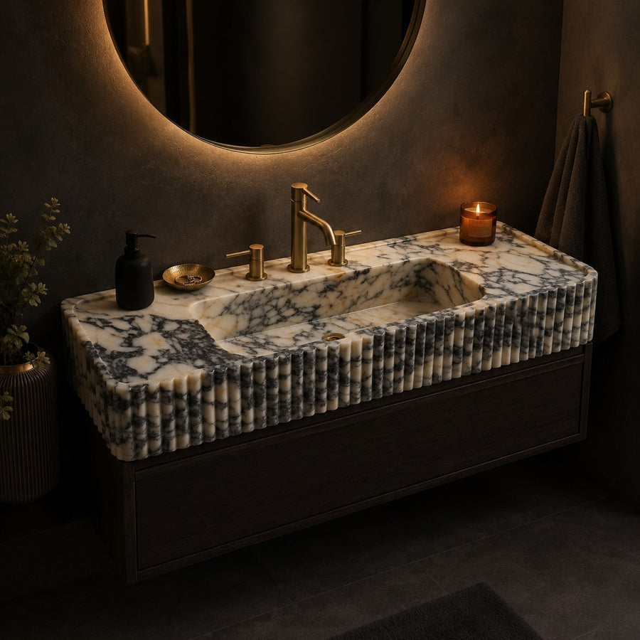 A floating bathroom vanity with a ribbed white and gray marble countertop, integrated sink, and brushed brass faucet and handles, shown in a dark modern bathroom setting with a lit candle and round wall mirror.