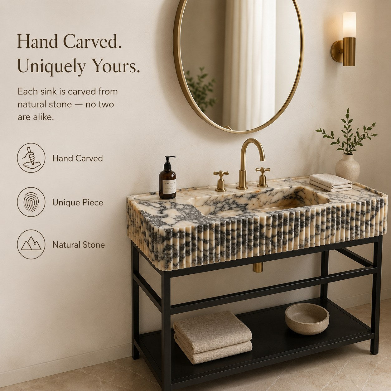 A freestanding rectangular bathroom sink with a carved natural stone top in beige, cream, and gray marbled tones, mounted on a black metal vanity. The sink is styled in a warm bathroom setting with a round gold-framed mirror, gold faucet, folded towels, a small plant, and a soap bottle.