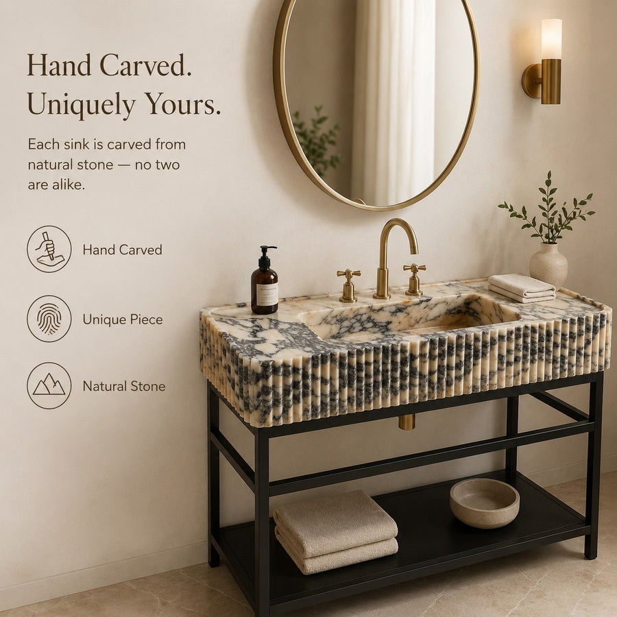 A freestanding rectangular bathroom sink with a carved natural stone top in beige, cream, and gray marbled tones, mounted on a black metal vanity. The sink is styled in a warm bathroom setting with a round gold-framed mirror, gold faucet, folded towels, a small plant, and a soap bottle.