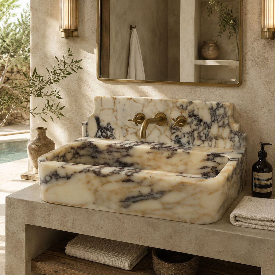 A rectangular marble vessel sink with cream, beige, and dark gray veining, paired with a matching marble backsplash and brass faucet, set in a luxurious bathroom with a concrete vanity and warm lighting.