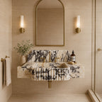 A modern wall-mounted bathroom sink with a cream and black marble pattern, paired with brushed gold fixtures, set against a beige stone wall with a matching arched mirror and wall sconces.