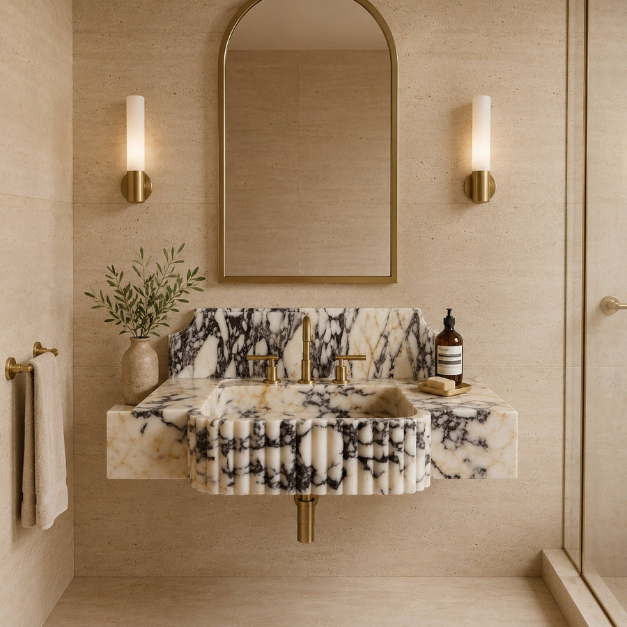 A modern wall-mounted bathroom sink with a cream and black marble pattern, paired with brushed gold fixtures, set against a beige stone wall with a matching arched mirror and wall sconces.