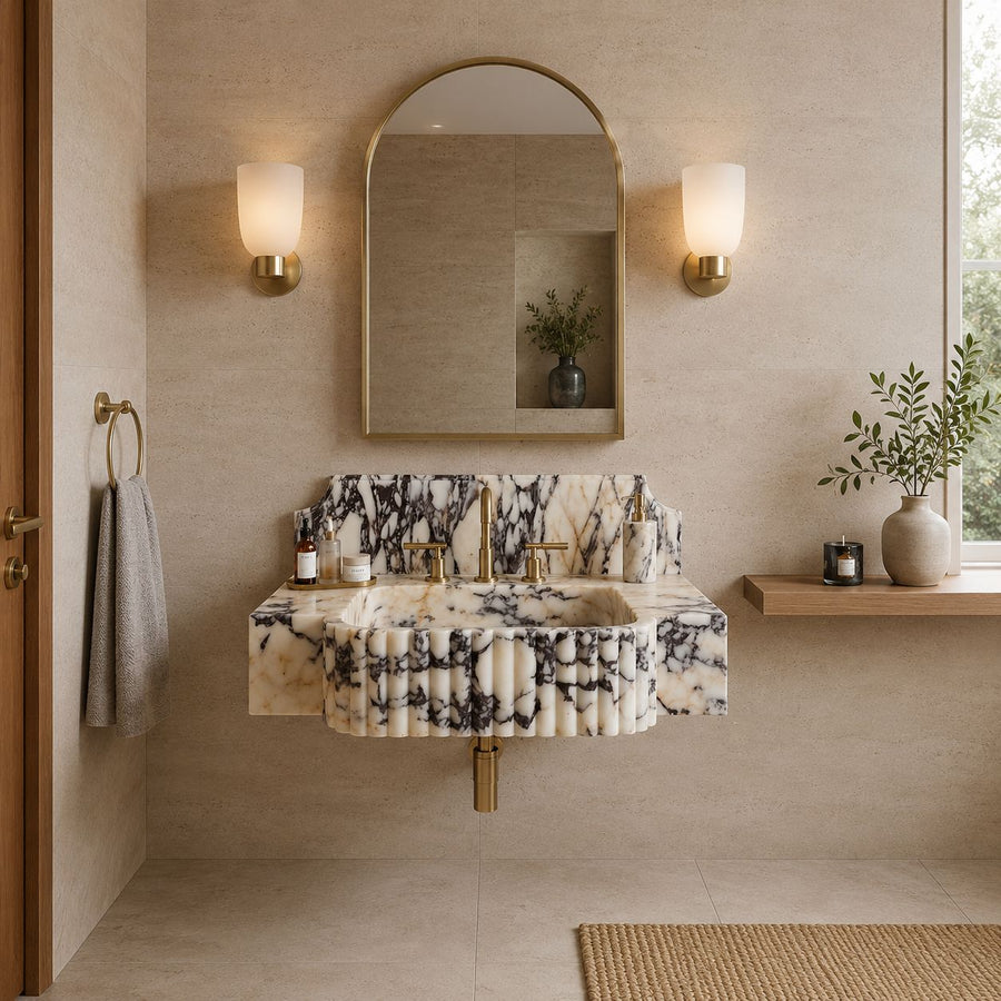 A floating vanity sink with a black-and-white marble pattern and pleated front, installed in a beige bathroom with a matching marble mirror, gold wall sconces, and a wooden shelf with a vase.