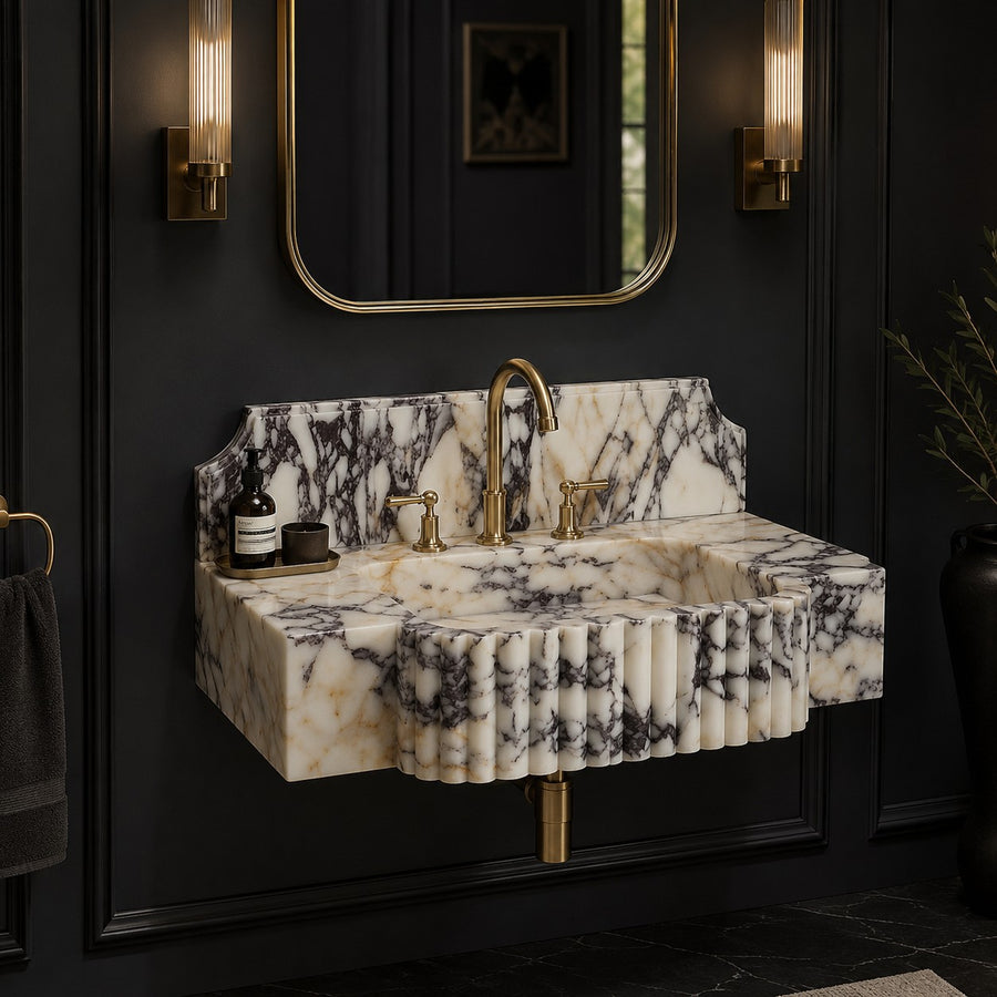 A wall-mounted marble sink with a fluted front edge, set into a dark paneled bathroom wall with a gold faucet, gold wall sconces, and a matching rectangular mirror.