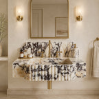 A luxury wall-mounted bathroom sink made of cream and black veined marble with pleated detailing, paired with a matching marble backsplash and gold-finished fixtures, set against a warm beige wall with a gold-framed mirror and wall sconces.