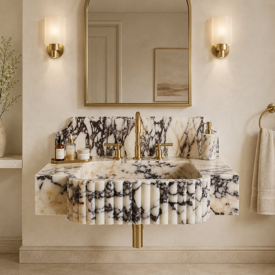 A luxury wall-mounted bathroom sink made of cream and black veined marble with pleated detailing, paired with a matching marble backsplash and gold-finished fixtures, set against a warm beige wall with a gold-framed mirror and wall sconces.