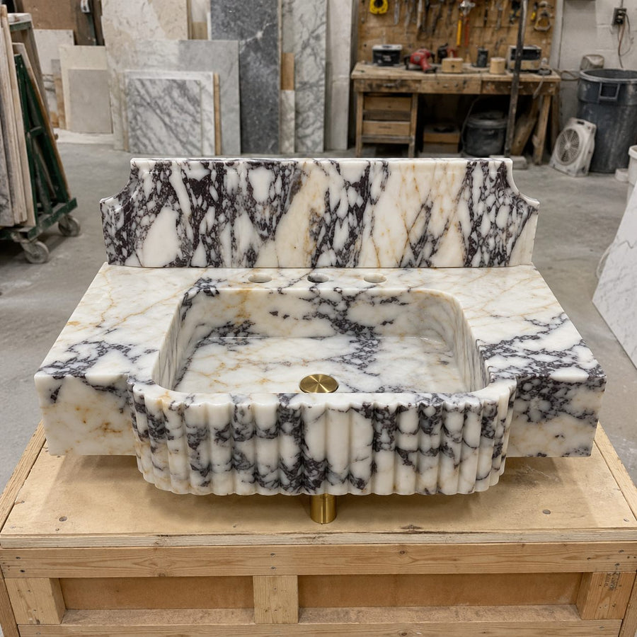 A freestanding rectangular marble sink basin with a fluted front edge and matching back panel, shown on a wooden display stand in a workshop. The stone has a white base with bold black and gray veining, and a small gold-toned drain is visible at the bottom center.