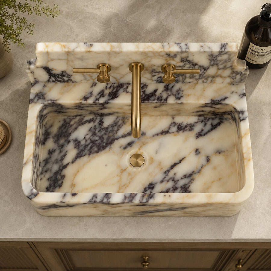 A rectangular farmhouse-style bathroom sink made of cream and gray marbled stone, paired with brushed brass faucet hardware and set into a light stone countertop.