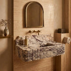 A floating bathroom sink made of fluted marble with a cream and gray veined pattern, mounted on a beige stone wall. The sink has a matching marble backsplash and a ribbed fluted front.