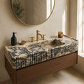 A floating bathroom vanity with a sculptural marble-look sink top and ribbed front, mounted above a wood cabinet. The scene is styled in a real bathroom with a round mirror, brass faucet and trim, a black soap dispenser, a small bowl, and a candle near a window.