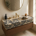 A floating bathroom vanity with a sculptural marble-look sink top and ribbed front, mounted above a wood cabinet. The scene is styled in a real bathroom with a round mirror, brass faucet and trim, a black soap dispenser, a small bowl, and a candle near a window.