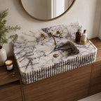 A rectangular white marble sink with dark veining and a ribbed fluted front, installed on a wooden vanity in a bathroom setting with a round mirror, brass faucet, and toiletries.