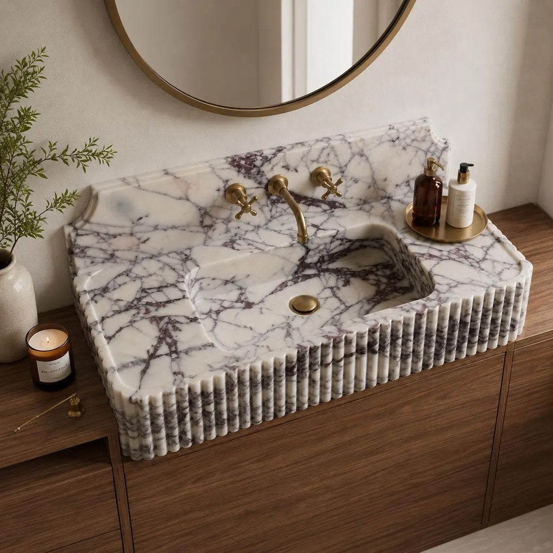 A rectangular white marble sink with dark veining and a ribbed fluted front, installed on a wooden vanity in a bathroom setting with a round mirror, brass faucet, and toiletries.