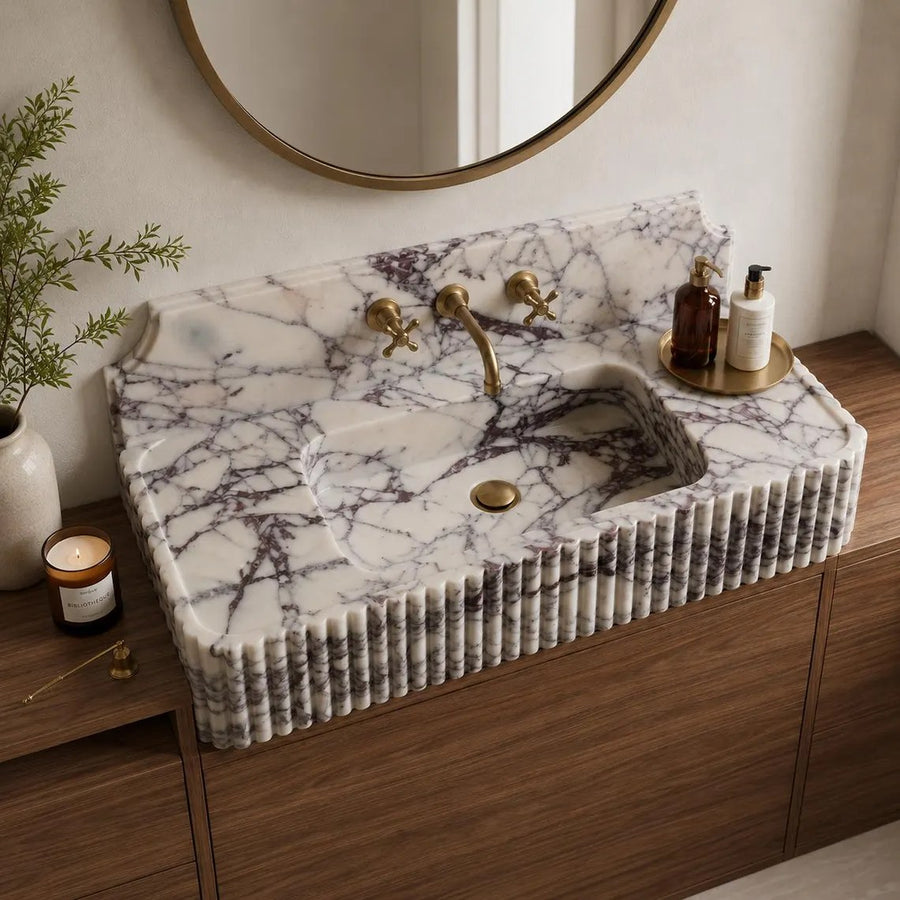 A rectangular white marble sink with dark veining and a ribbed fluted front, installed on a wooden vanity in a bathroom setting with a round mirror, brass faucet, and toiletries.