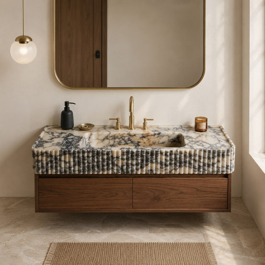 A floating bathroom vanity with a polished marble countertop and matching marble sink surround, paired with a dark wood cabinet and gold-tone faucet. The vanity is shown in a styled bathroom setting with a large gold-framed mirror, a hanging globe light, a soap dispenser, and a window light casting soft shadows.