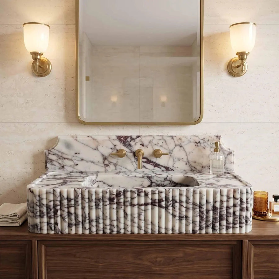 A luxury bathroom vanity featuring a rectangular marble sink with a fluted, ribbed front and a matching marble backsplash. The sink is mounted on a dark wood cabinet, flanked by a large rectangular mirror with a gold frame and two matching gold wall sconces on the beige stone wall.