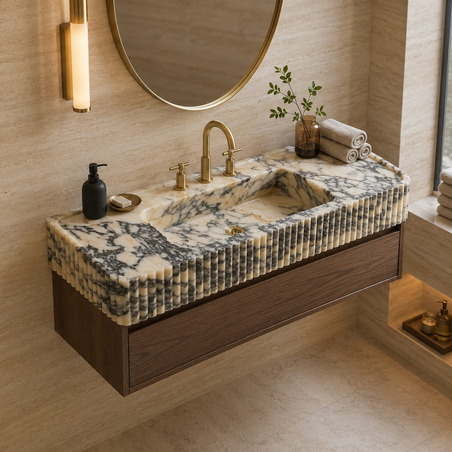 A floating bathroom vanity with a carved stone sink top in cream and gray marbled stone, a dark wood cabinet, a gold faucet, and matching gold wall hardware in a warm beige bathroom setting.
