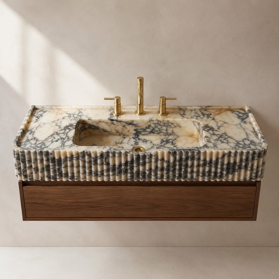A floating bathroom vanity with a rectangular carved stone sink top in cream and gray marbled stone, a ribbed fluted front, and a dark wood base, shown with a matte brass faucet set against a warm beige wall.