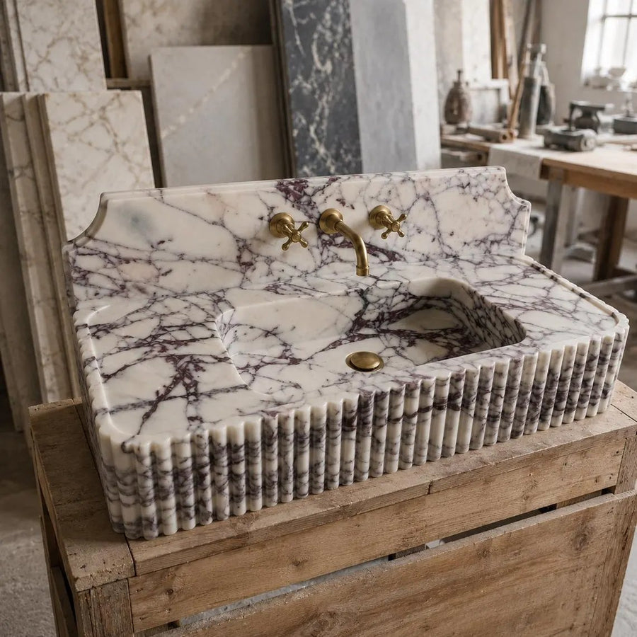 A freestanding rectangular marble sink with a ribbed fluted base, featuring a white and gray veined pattern with dark purple-gray streaks, set on a rustic wooden crate in a workshop environment.