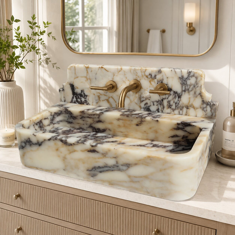 A rectangular marble vessel sink with a matching marble backsplash and gold faucet, set on a light wood vanity in a bright bathroom.