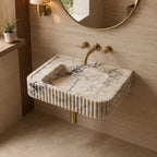 A modern wall-mounted bathroom sink made of cream and gray veined marble with fluted edges, paired with brushed gold fixtures and a round mirror on a beige stone wall.