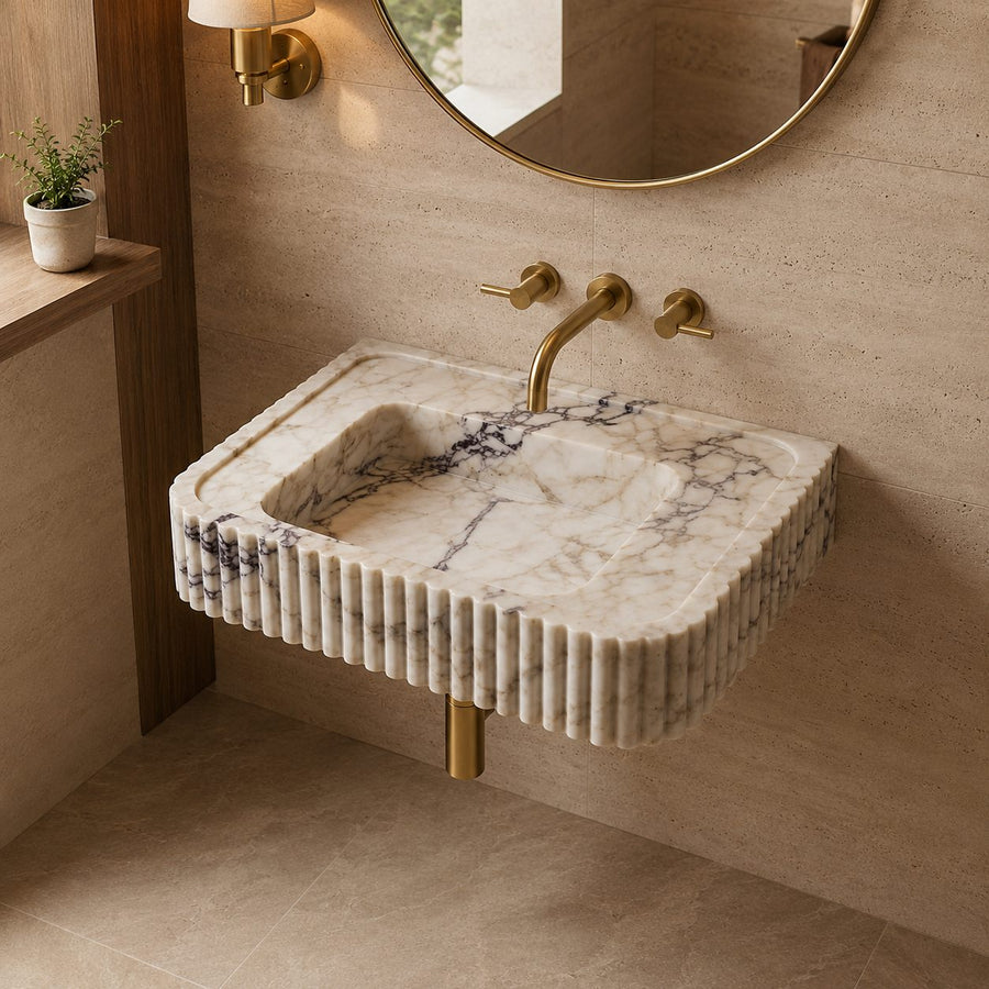 A modern wall-mounted bathroom sink made of cream and gray veined marble with fluted edges, paired with brushed gold fixtures and a round mirror on a beige stone wall.