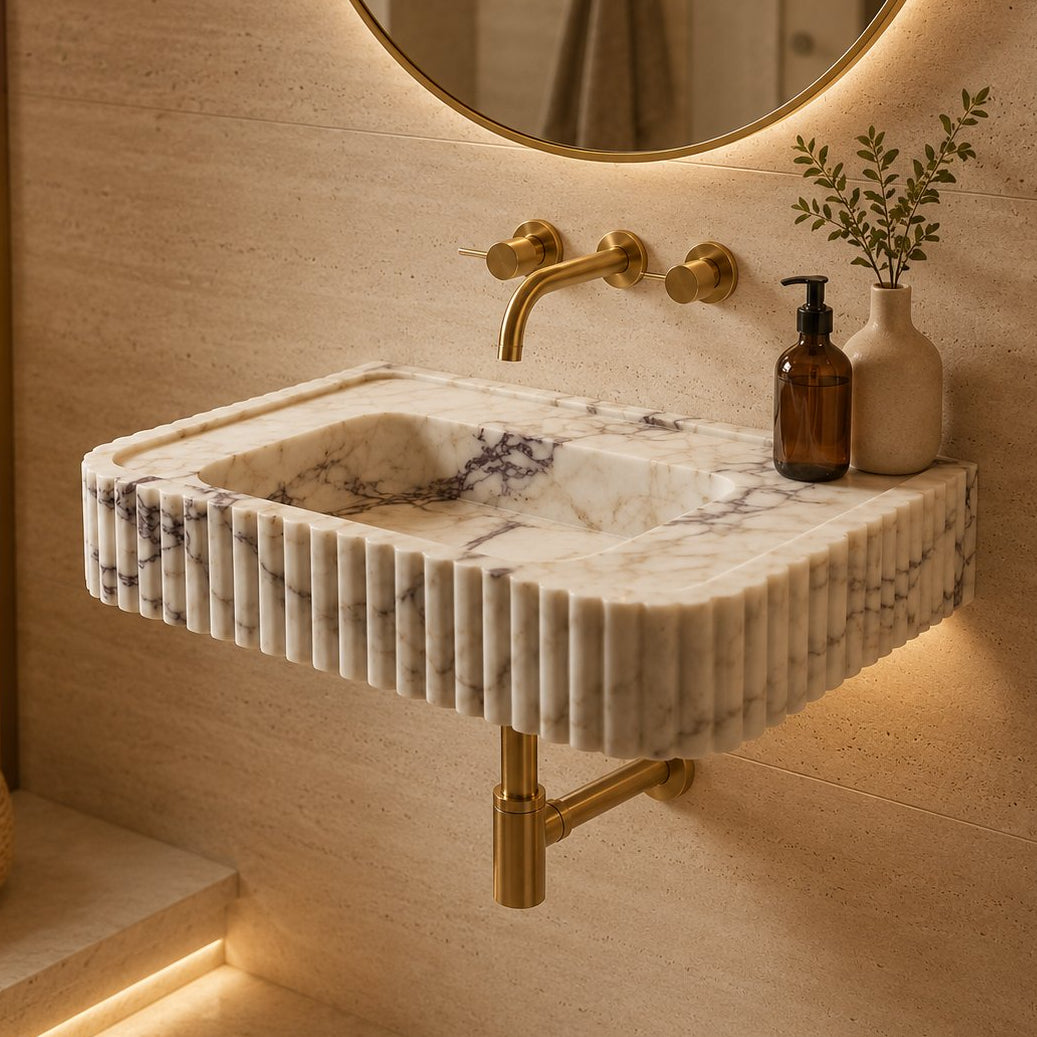 A modern wall-mounted bathroom sink made of fluted white marble with dark veining, paired with brushed brass faucets and fixtures, set against a warm beige stone wall with a lit round mirror.