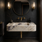 A modern wall-mounted bathroom sink with a ribbed cream marble front and dark veining, paired with brushed gold fixtures, a round mirror, and wall sconces against a dark charcoal wall.