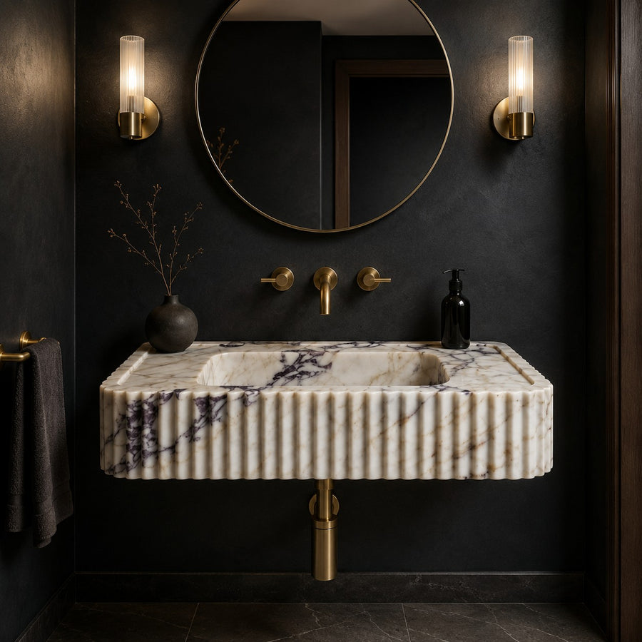 A modern wall-mounted bathroom sink with a ribbed cream marble front and dark veining, paired with brushed gold fixtures, a round mirror, and wall sconces against a dark charcoal wall.