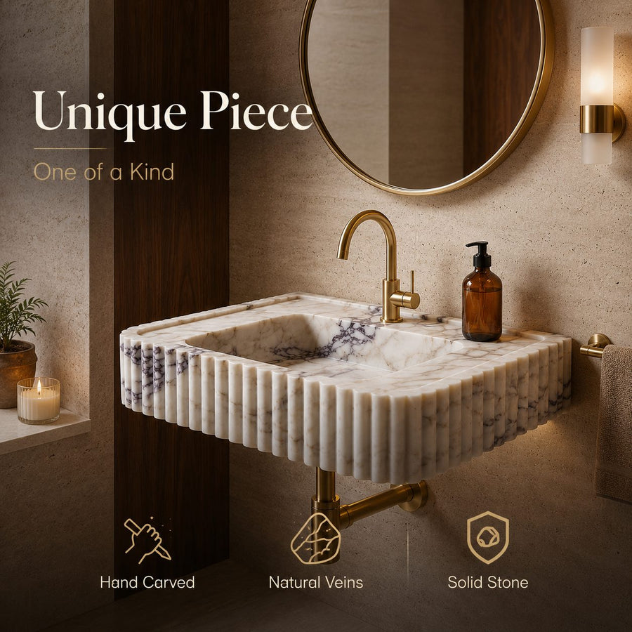 A wall-mounted rectangular stone sink with a fluted front, white marble-like surface, and dark veining, shown in a bathroom setting with a gold faucet, round mirror, and warm ambient lighting. Text on the image reads 'Unique Piece One of a Kind' with feature icons below.