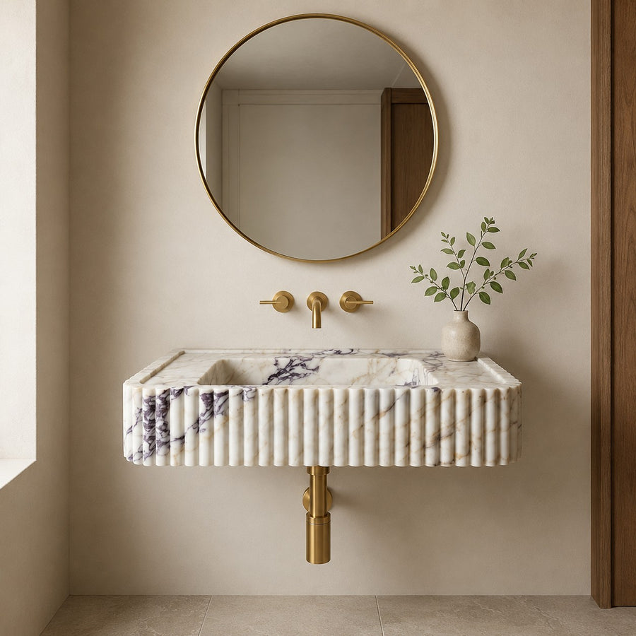 A modern wall-mounted sink with a ribbed white marble front, a round gold-framed mirror above, and gold wall-mounted fixtures in a warm beige bathroom setting.