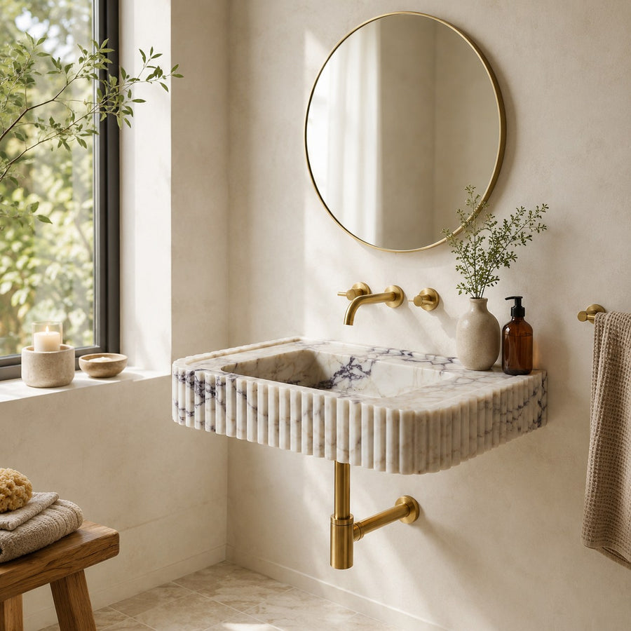 A modern wall-mounted sink with a ribbed cream and blue-gray marble pattern, paired with brushed gold fixtures, a round gold-framed mirror, and a neutral bathroom setting.