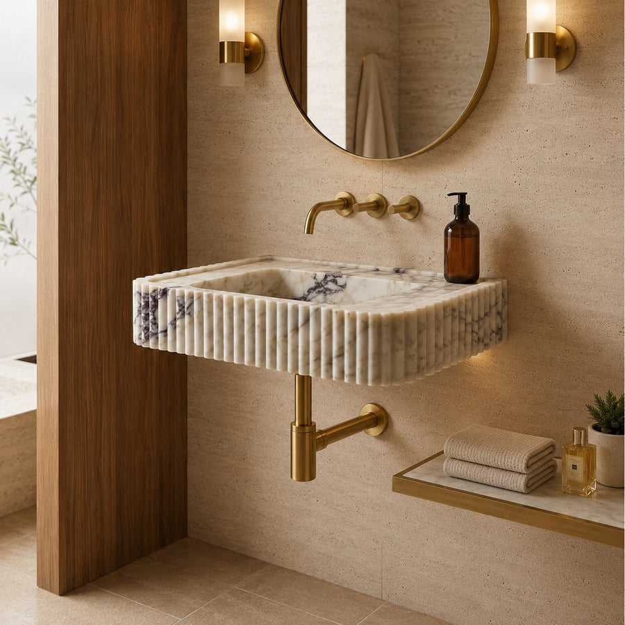 A modern wall-mounted bathroom sink made of ribbed cream marble with subtle blue-gray veining, paired with brushed gold hardware, set in a warm beige bathroom with a round mirror and sconces.