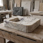 A rectangular stone basin with fluted sides and a recessed center, made from light beige marble with dark veining, displayed on a rustic wooden workbench in a stone workshop.