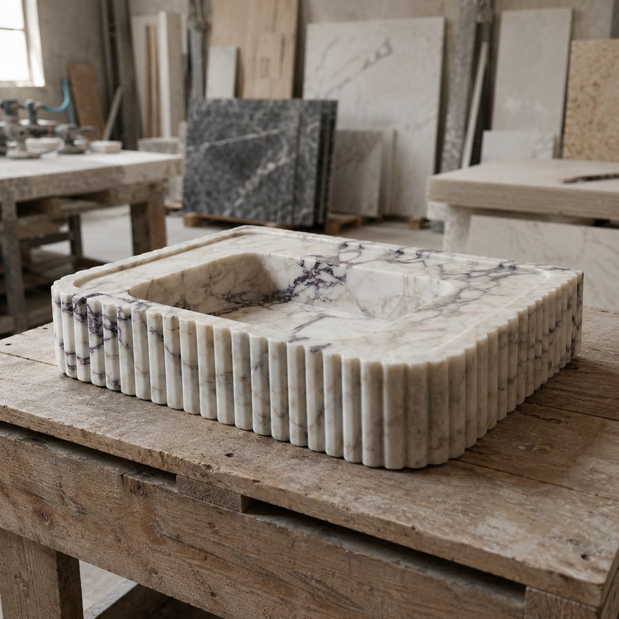 A rectangular stone basin with fluted sides and a recessed center, made from light beige marble with dark veining, displayed on a rustic wooden workbench in a stone workshop.