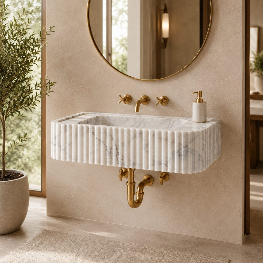 A modern wall-mounted bathroom sink made of white fluted marble with subtle gray veining, paired with brushed gold hardware and a round gold-framed mirror on a beige stone wall.