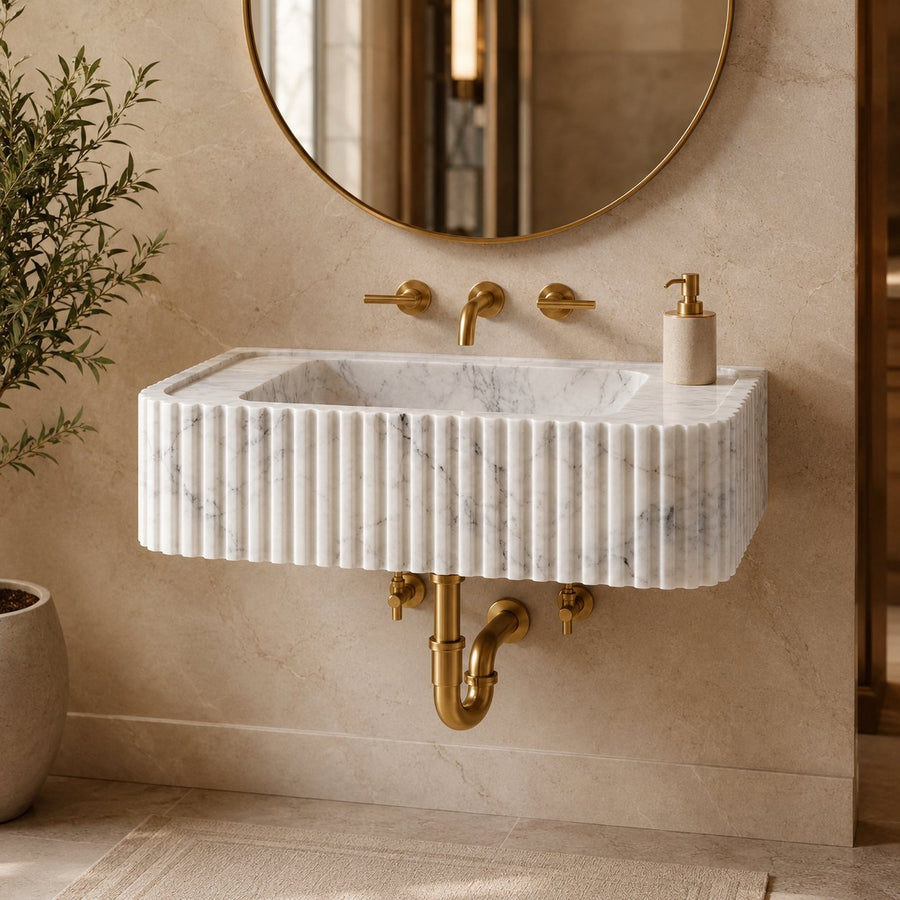A modern wall-mounted rectangular sink made of white fluted marble with subtle gray veining, paired with brushed gold fixtures, a round gold-framed mirror, and a beige tiled bathroom wall.