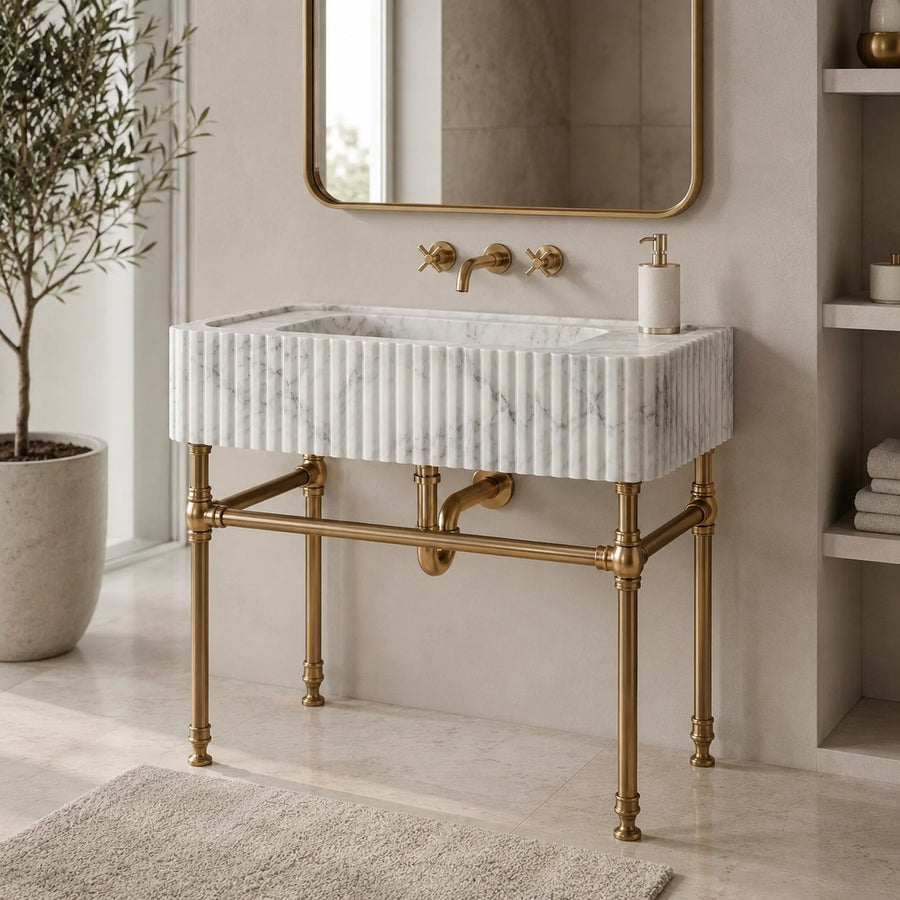 A freestanding rectangular bathroom sink with a white marble-look ribbed front, supported by ornate brass legs and matching brass faucet hardware, shown in a styled bathroom setting with a mirror, plant, and shelving.