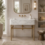 A modern bathroom vanity featuring a white marble sink top with vertical fluting, supported by ornate brass legs, set against a beige wall with a gold-framed mirror and brass fixtures.
