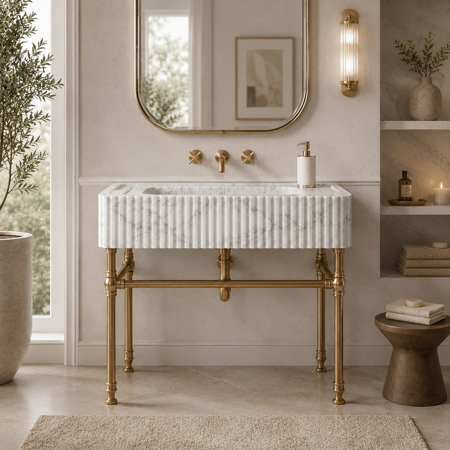 A modern bathroom vanity featuring a white marble sink top with vertical fluting, supported by ornate brass legs, set against a beige wall with a gold-framed mirror and brass fixtures.